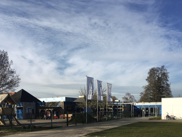 Großes Schulgebäude aus Backsteinen mit einem grasbewachsenen Vorgarten, umgeben von einem Metallzaun, mit einem Weg, Bäumen, Fahnen, einem geparkten Fahrrad, nahegelegenen Häusern und einem bewölkten Himmel.