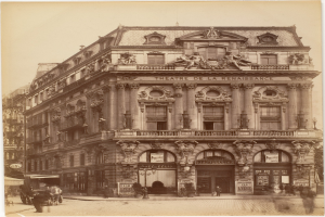 Schwarz-weiß-Foto des Théâtre de la Renaissance in Paris, ein mehrstöckiges Gebäude mit Rundbogenfenstern, Säulen und skulpturalen Verzierungen an der Fassade, mit Passanten und Pferdekutschen auf der Straße davor und Himmel darüber.