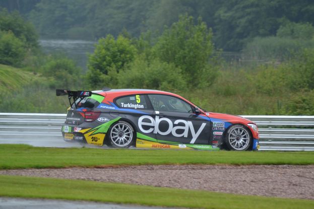 Ein Auto auf einer Rennstrecke fahrend, das Rauch ausstößt, umgeben von Gras, einem Zaun, Pflanzen, Bäumen und Wasser, mit sichtbarem Text und einem Rad am Auto.