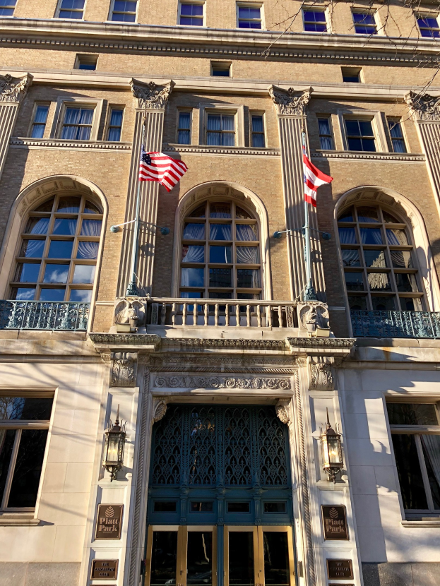 Außenansicht des Plaza Hotels in New York City, mit Glasfenstern, Türen, Säulen, Geländern, Leuchten, Schildern und Flaggen vor einem Himmel als Hintergrund.