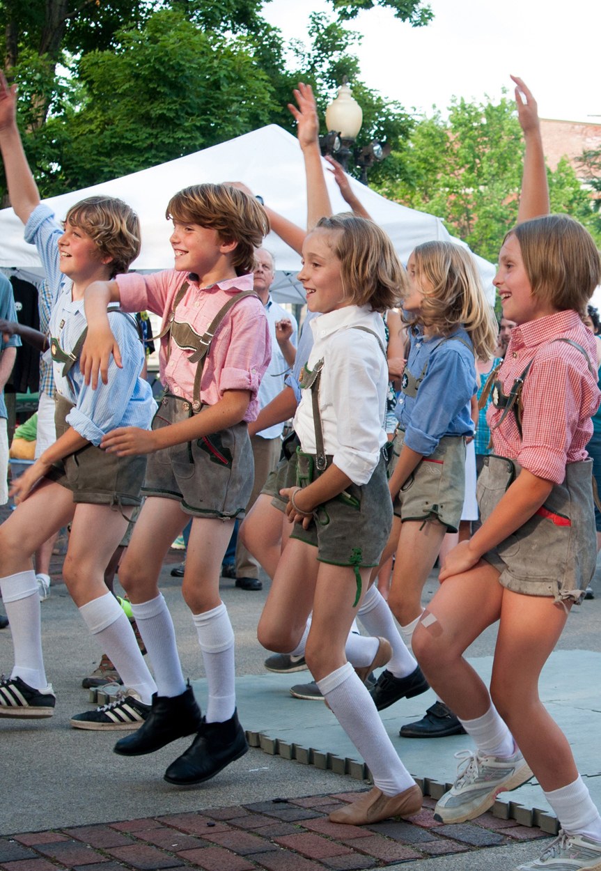 Kinder in traditioneller bayrischer Tracht, die auf einer Straße tanzen, umgeben von einer Menge, Zelten, Laternenmasten, Bäumen, Gebäuden und einem klaren blauen Himmel.
