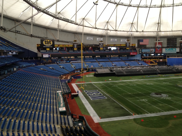 Großes Stadioninnere mit einem Fußballfeld, Sitzreihen auf beiden Seiten, Hintergrundtafeln mit Text und Fahnen sowie Dachkonstruktion mit Beleuchtung und Stützstangen.