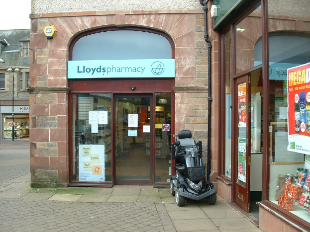 Eine Apothekenfiliale mit einem vor ihr geparkten Fahrzeug und einem Gebäude in der linken Ecke.