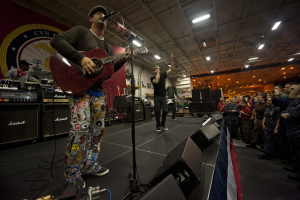 Ein Mann, der als Chris Stapleton identifiziert wird, spielt eine akustische Gitarre auf der Bühne, trägt eine Mütze, hat ein Mikrofon, Lautsprecher, eine Flagge und andere Instrumente in der Nähe, während eine Menge unter Deckenlampen mit einem Banner hinter ihm zusieht.