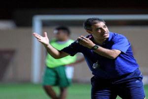 Ein Mann in einem blauen T-Shirt und Hose steht auf einem Fußballfeld und hält ein Mikrofon in der Hand, während er auf seine Spieler zeigt. Im Hintergrund ist eine Person in einem grünen Kleid, ein Tor und eine Wand zu sehen.