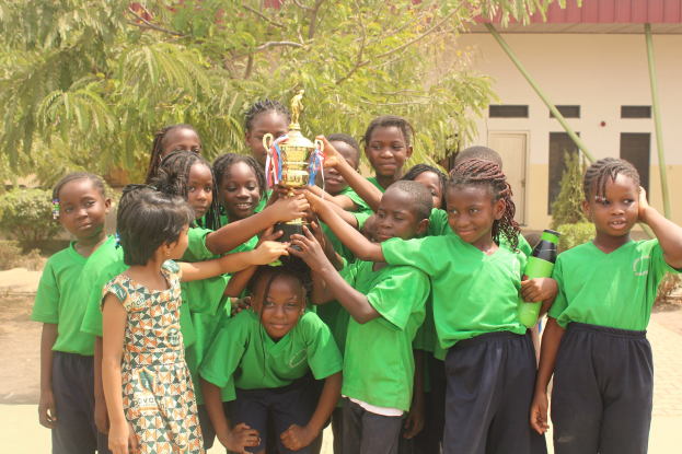 Gruppe von Kindern mit Pokal in einem Schulhof mit Bäumen und einem Gebäude im Hintergrund