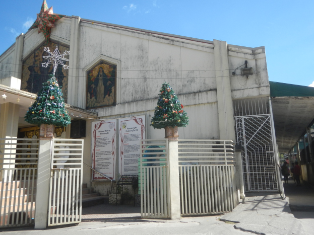 Kirche mit zwei geschmückten Weihnachtsbäumen am Eingang, Treppe hinauf zum Gebäude, einige Menschen herumlaufend und ein bewölkter Himmel im Hintergrund.