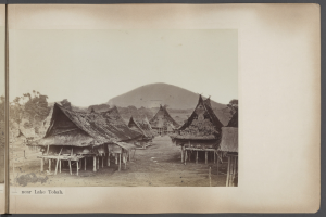 Schwarzes und weißes Foto eines Dorfes mit Hütten, Bäumen und einem Berg im Hintergrund, mit Text unten.