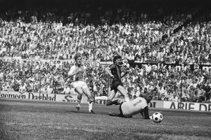 Schwarzes Foto von Füßballspielern auf einem Feld während eines Bundesliga-Spiels zwischen Ajax und Hannover 96 in der Saison 1978-79, mit Zuschauern im Hintergrund.