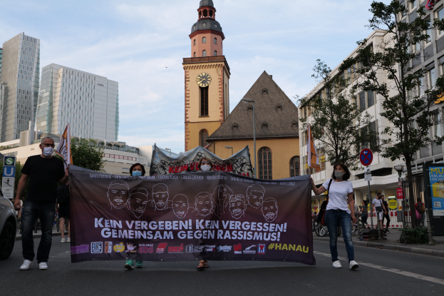 Eine Gruppe von Menschen in Masken marschiert auf einer Straße und hält ein Banner hoch, mit einem geparkten Auto auf der linken Seite, Gebäuden, Bäumen, Schildern, Pfosten, einem Kirchturm und einem klaren blauen Himmel im Hintergrund.