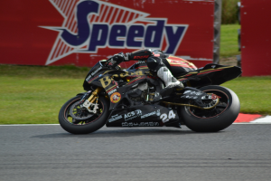 Eine Person in Helm und Schuhwerk fährt ein Motorrad auf einer Rennstrecke, die von Gras umgeben ist, mit einer Tafel im Hintergrund, auf der Text zu sehen ist.