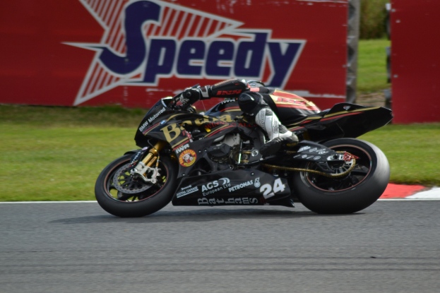 Eine Person in Helm und Schuhwerk fährt ein Motorrad auf einer Rennstrecke, die von Gras umgeben ist, mit einer Tafel im Hintergrund, auf der Text zu sehen ist.