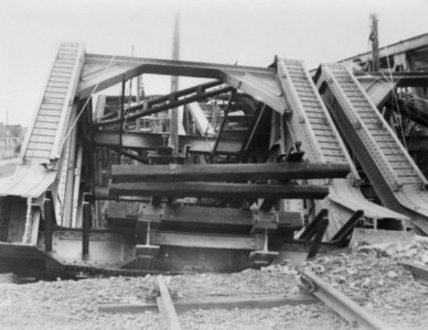 Ein Schwarz-Weiß-Foto einer Bahnschiene, die zu einer baufälligen Brücke führt, umgeben von Gebäuden unter einem klaren Himmel.