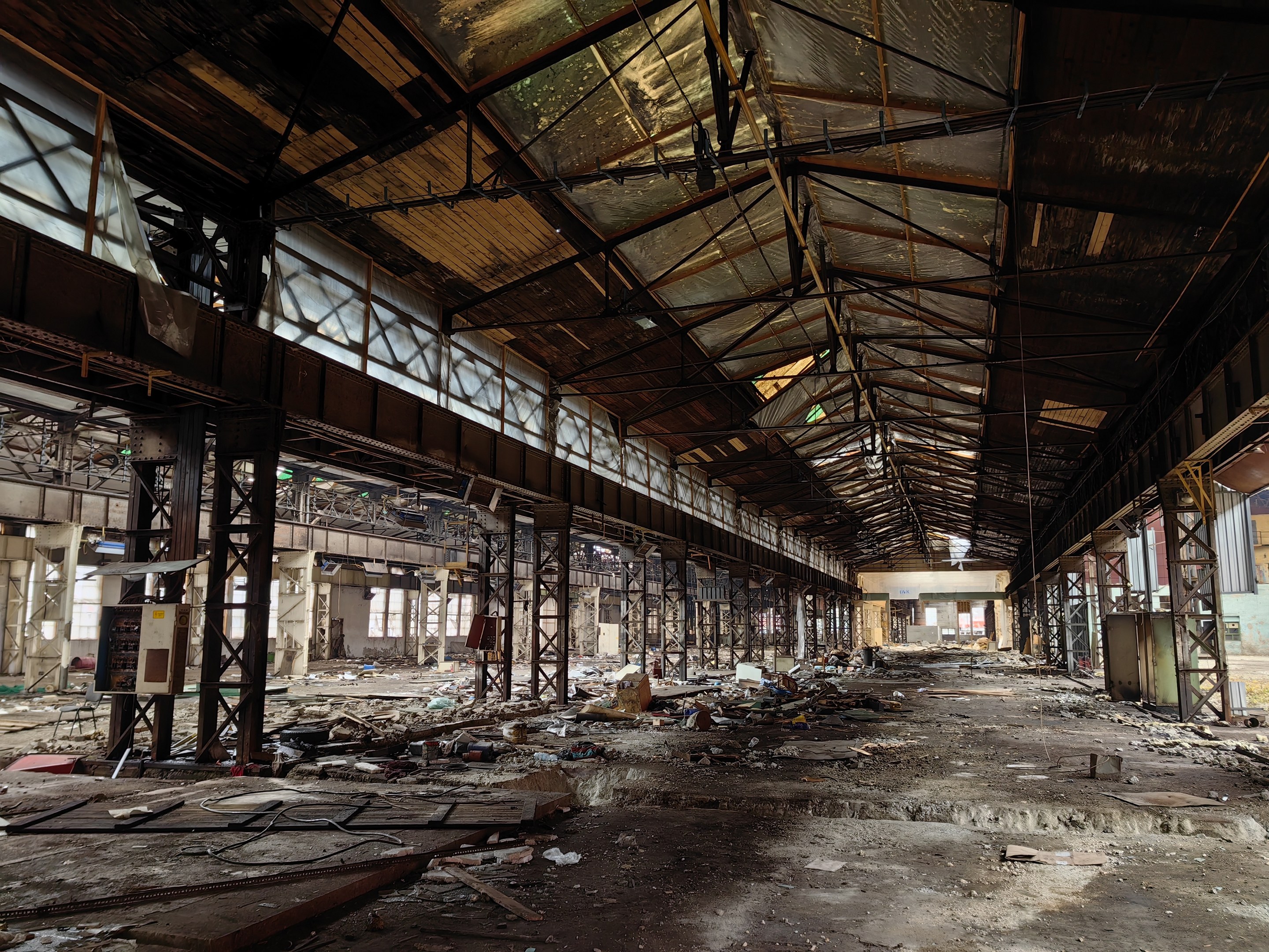 Verlassene Fabrikhalle mit verstreutem Schutt, zerbrochenen Fenstern, Metallstangen, Deckenlampen und einem beschädigten Dach.