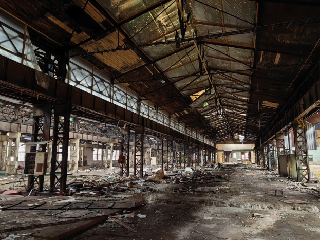 Verlassene Fabrikhalle mit verstreutem Schutt, zerbrochenen Fenstern, Metallstangen, Deckenlampen und einem beschädigten Dach.