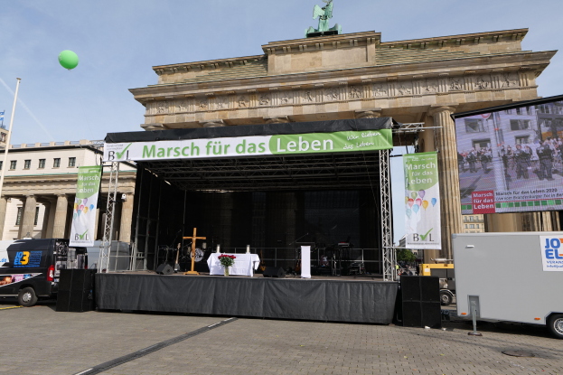 Bühne vor dem Brandenburger Tor mit einem Tisch, Bannern, Lautsprechern, Fahrzeugen, Gebäuden, einer Statue, einer Flagge und Wolken im Hintergrund.