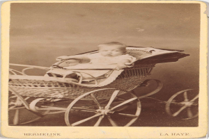 Ein altes Schwarz-Weiß-Foto eines Babys in einem hölzernen Kinderwagen, das ein weißes Strampler und einen friedlichen Ausdruck trägt, mit kursiver Schrift am unteren Rand.