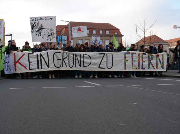 Gruppe von Menschen mit einem Transparent mit der Aufschrift "Kein Grund zu Feiern" bei einer Demonstration gegen die Sparpolitik der deutschen Regierung, mit Gebäuden, Bäumen und einem klaren Himmel im Hintergrund.