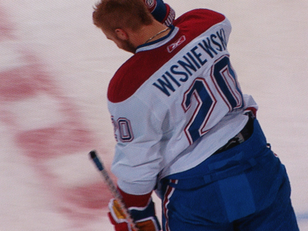 Ein Eishockeyspieler in einem "Wisniewski"-Trikot steht auf dem Eis, hält einen Eishockeyschläger und hat einen konzentrierten und entschlossenen Gesichtsausdruck.