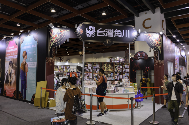 Eine Gruppe von Menschen steht vor einem Messestand, einige halten Taschen, mit Bändern, Informationsplakaten, Deckenbeleuchtung und Displayregalen im Hintergrund, wahrscheinlich für die Tokyo Anime Expo 2019.