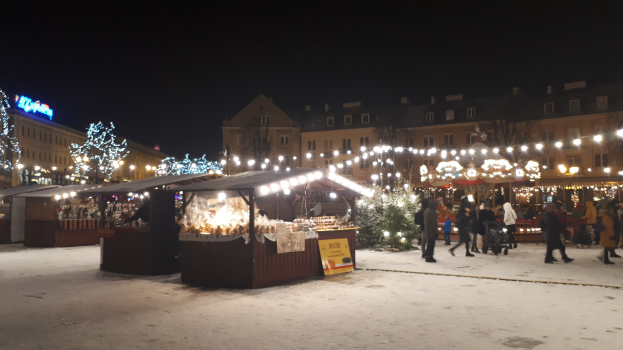 Ein verschneiter Weihnachtsmarkt bei Nacht mit Menschen, Buden, Pflanzen und Gebäuden unter einem bewölkten Himmel.