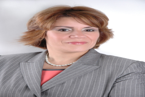 Eine professionelle Frau in einem marineblauen Anzug mit einem weißen Hemd und einer gemusterten Krawatte steht selbstbewusst gegen einen weißen Hintergrund, ihre Haltung aufrecht und ihr Blick direkt.