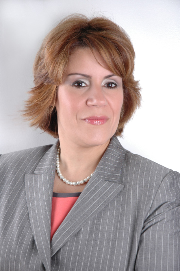 Eine professionelle Frau in einem marineblauen Anzug mit einem weißen Hemd und einer gemusterten Krawatte steht selbstbewusst gegen einen weißen Hintergrund, ihre Haltung aufrecht und ihr Blick direkt.