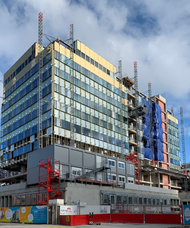 Großes Gebäude im Bau in einer Stadt mit Glasfenstern, Metallrahmen, Treppe, Umzäunung, Schild, Versorgungsmasten, Straßenlaterne, nahen Bäumen und bewölktem Himmel.