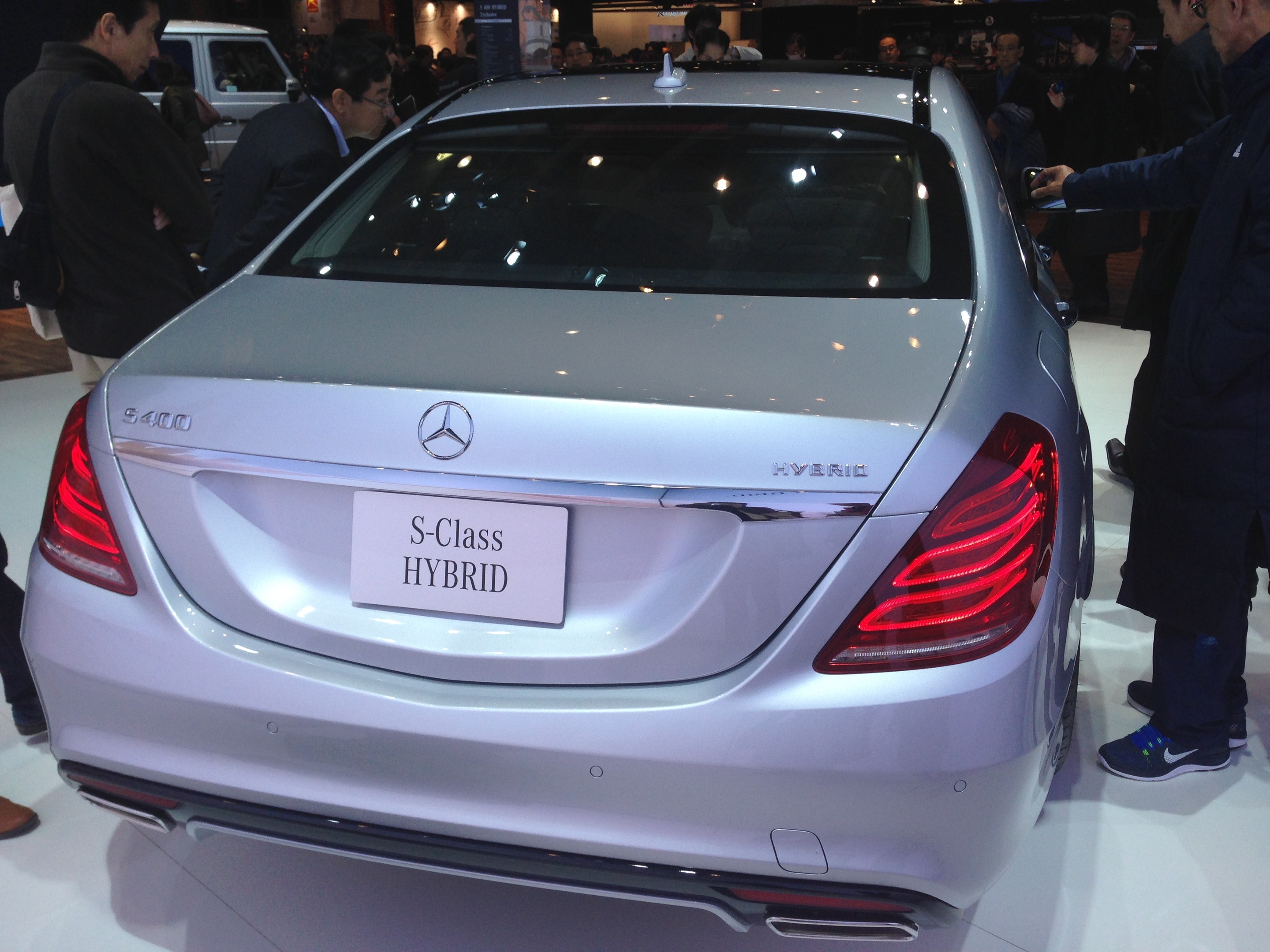 Silberner Mercedes S-Klasse Hybrid auf einer Automesse präsentiert, umgeben von Menschen in verschiedenen farbigen Kleidern, mit anderen Fahrzeugen und Beleuchtung im Hintergrund.