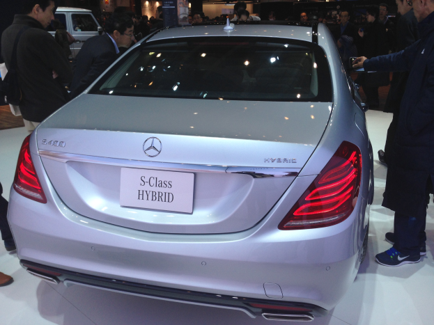 Silberner Mercedes S-Klasse Hybrid auf einer Automesse präsentiert, umgeben von Menschen in verschiedenen farbigen Kleidern, mit anderen Fahrzeugen und Beleuchtung im Hintergrund.