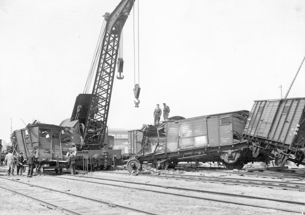Schwarzes und weißes Bild eines Zugunglücks mit einem Kran, der einen Zugwagen auf die Schienen hebt, mehrere Menschen in der Nähe und im Hintergrund Gebäude unter einem klaren Himmel zu sehen.