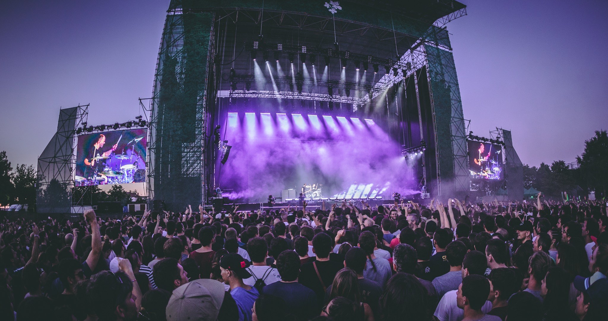 Eine gro├če Menge bei einem Musikfestival schaut auf einer hell erleuchteten Bühne mit Instrumenten und Bildschirmen, die vor einem Geb├Ąude, B├Ąumen und einem klaren blauen Himmel stehen.