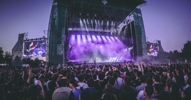 Eine gro├če Menge bei einem Musikfestival schaut auf einer hell erleuchteten Bühne mit Instrumenten und Bildschirmen, die vor einem Geb├Ąude, B├Ąumen und einem klaren blauen Himmel stehen.
