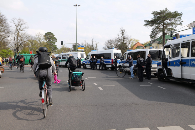 Eine Gruppe von Menschen, die auf Fahrrädern eine Straße entlangfahren, mit Polizeiwagen, Fahrzeugen, Bäumen und Gebäuden im Hintergrund.