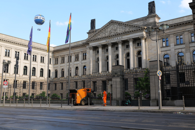 Großes modernes Regierungsgebäude mit vielen Fenstern, umgeben von Straßenmöbeln und Fußgängern, mit wehenden Flaggen davor.