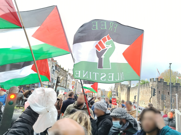 Eine Gruppe von Menschen mit palästinensischen Flaggen vor einer Menge, im Hintergrund Gebäude, Bäume, Pfosten, Verkehrszeichen, Schilder und eine Wand, die sich versammelt haben, um gegen die israelische Besetzung Palästinas zu protestieren.