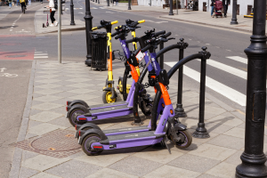 Eine Reihe von E-Scootern, die entlang einer Straße mit Fußgängern, Fahrrädern, einem Müllcontainer und Gebäuden im Hintergrund geparkt sind.