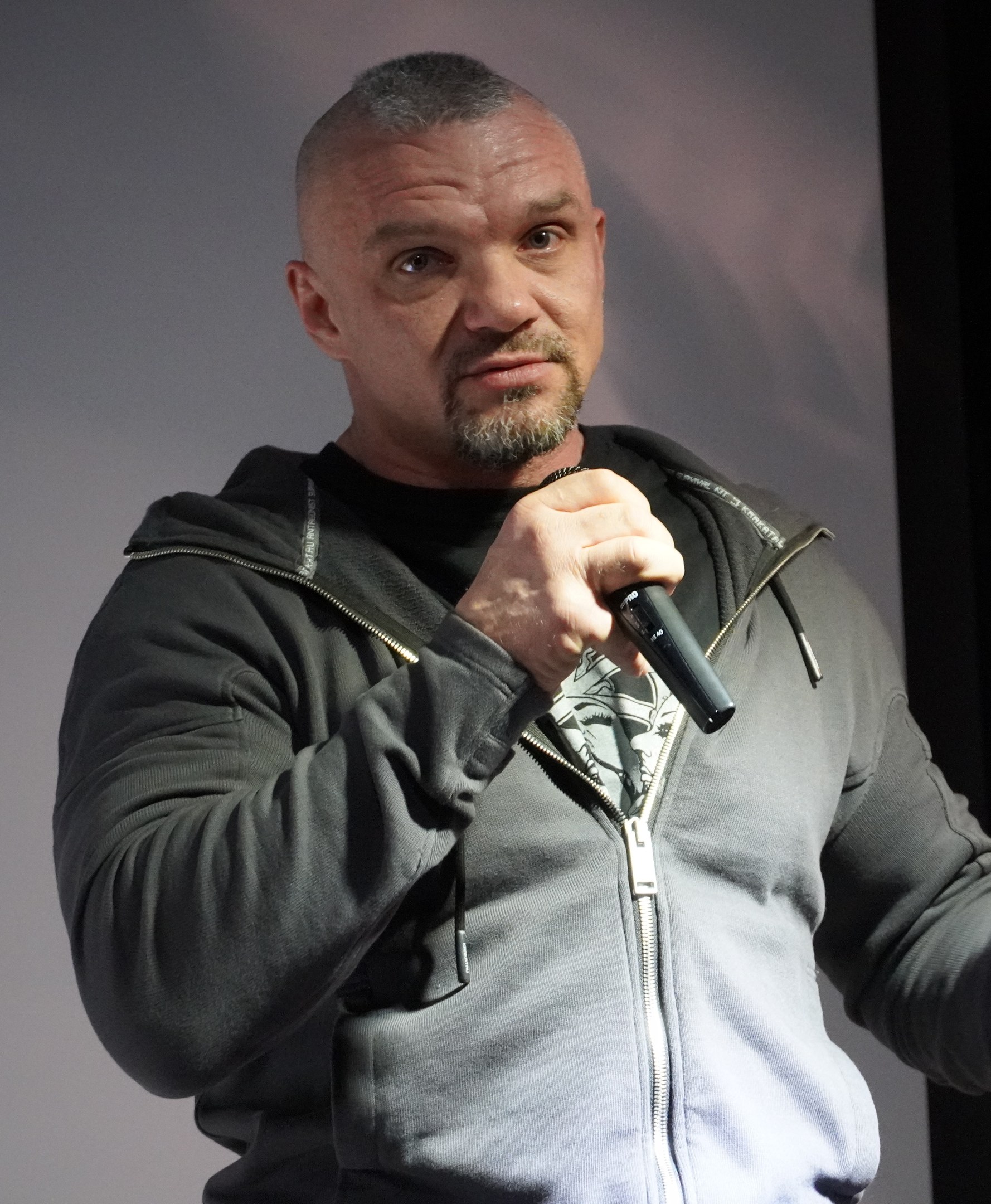 Ein Mann in einer schwarzen Kapuzenjacke und einem schwarzen Anzug mit weißem Hemd, der ein Mikrofon hält und selbstbewusst vor einer Wand steht.