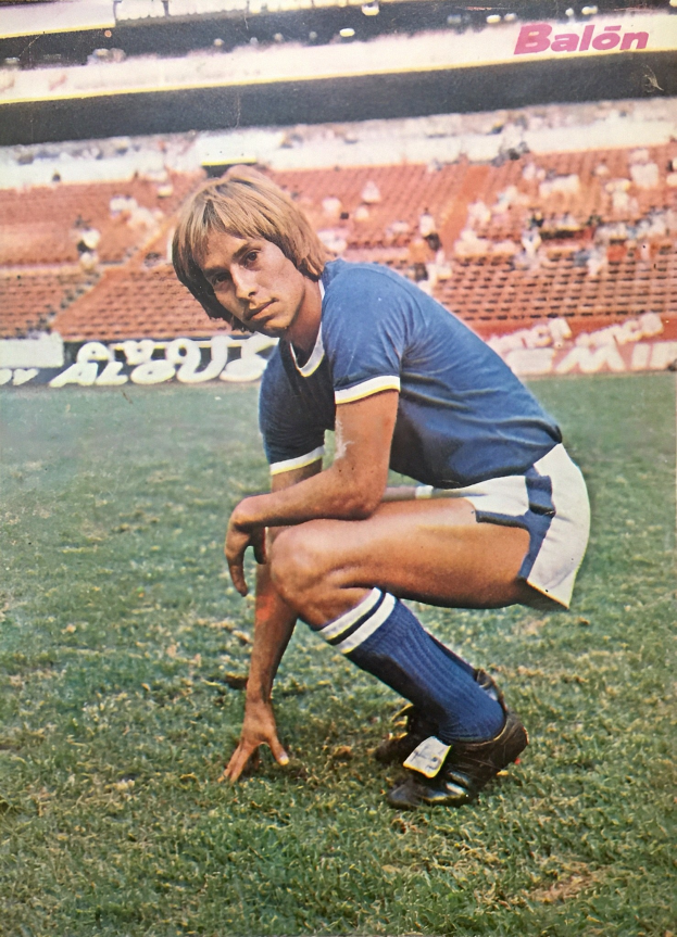 Ein Mann kniet auf einem Fußballfeld umgeben von sitzenden Personen mit Schildern, trägt Sportschuhe.