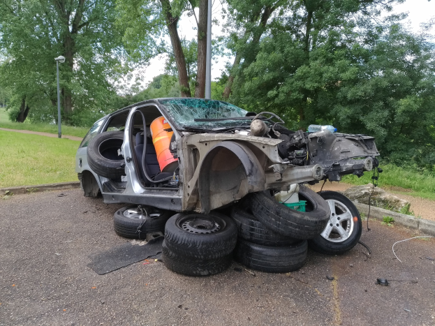 Ein umgestürzter Wagen am Straßenrand mit verstreuter Trümmer, umgeben von Gras, Pflanzen, Bäumen, einem Straßenpfahl und einem bewölkten Himmel.