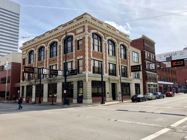 Ein großes Backsteingebäude, das früher das Rathaus war, steht an der Ecke einer belebten Stadtstraße, umgeben von anderen Gebäuden, Verkehr, Passanten und Bäumen unter einem bewölkten Himmel.