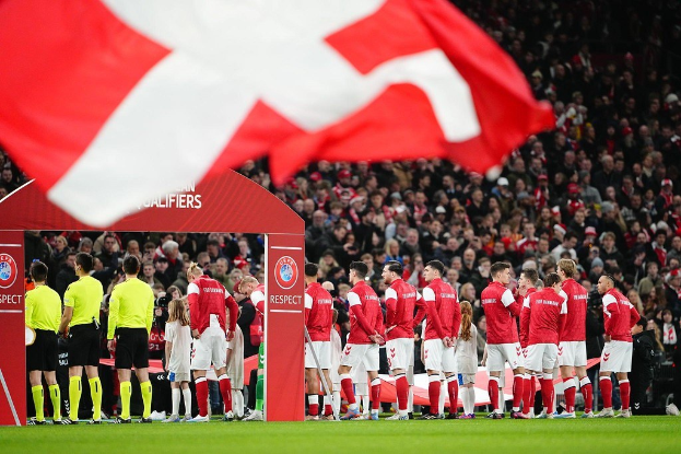 Eine Gruppe von Menschen auf einem Fußballfeld mit einer roten und weißen Fahne im Vordergrund, einem Bogen mit der Aufschrift "Bayern München vs. Bayern München Wett-Tipps & Vorschau" im Hintergrund und einer großen Menge im Stadion.