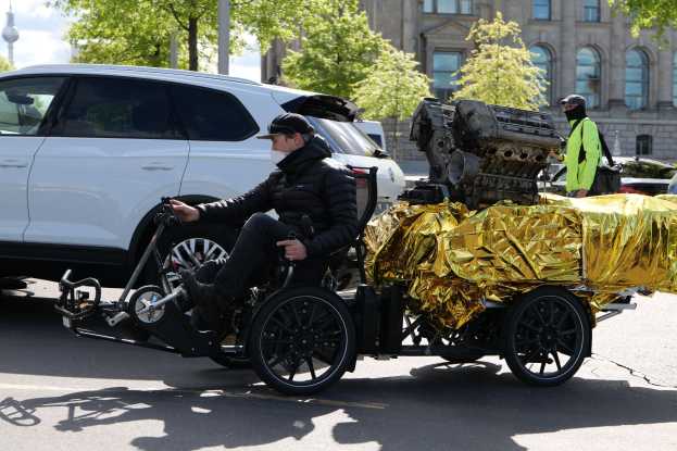 Ein Mann im Rollstuhl mit einem großen Motor an dessen Rückseite, umgeben von Fahrzeugen auf einer Straße mit Bäumen, Gebäuden und einem klaren blauen Himmel im Hintergrund, trägt eine schwarze Jacke und eine Kappe und hält ein Objekt.