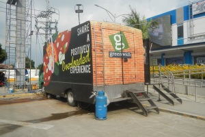 Lastwagen vor einem Gebäude mit einer Tafel, auf der "Teilen Sie eine positiv überladene Erfahrung" steht, mit Stufen, einem Zylinder auf dem Boden, Geländern, Strommasten mit Drähten, Laternenpfählen, Bäumen und einem bewölkten Himmel.