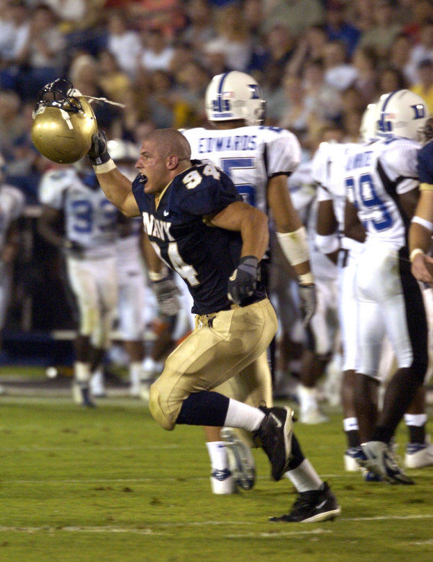Ein Fußballspieler in dunkler Uniform rennt mit dem Ball, umgeben von Teamkollegen in Sportkleidung und Helmen, während Zuschauer in Stühlen zusehen.
