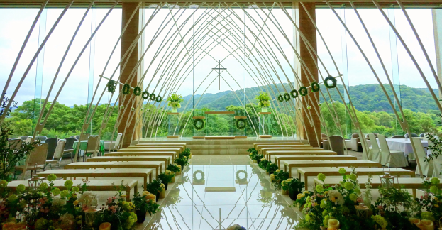 Eine Hochzeitszeremonie in einem großen, geschmückten Saal mit Tischen in Halbkreisform, Blumenbouquets und einer Aussicht auf Bäume, Hügel und einen klaren blauen Himmel im Hintergrund.